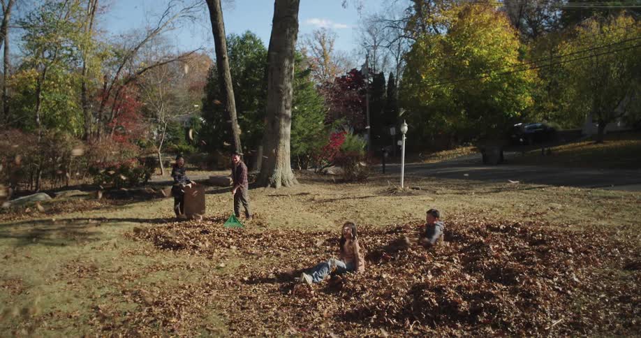 Two children lying on dry leaves in a garden and the father with a shovel throwing other leaves