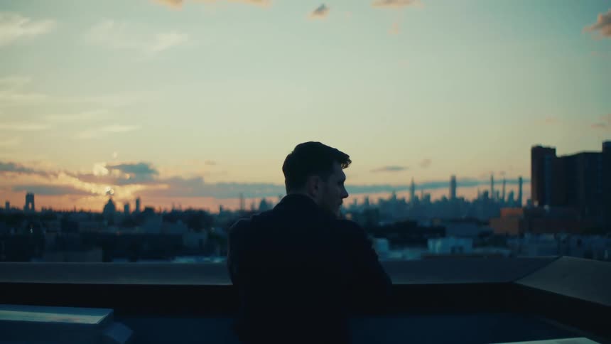 A man is standing on a rooftop at sunset overlooking a city skyline with tall buildings and a colorful sky