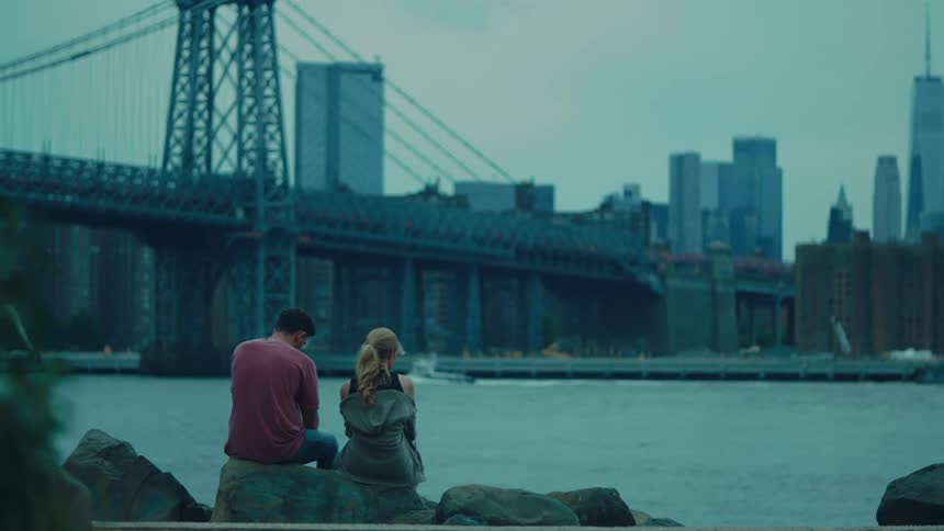 Man and a woman sitting side by side on a rock by the sea with a metropolis in the background