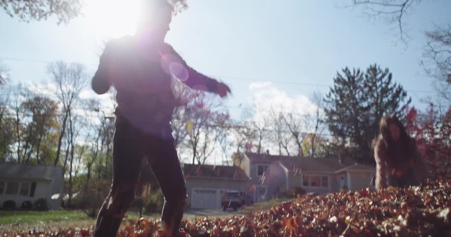 Two children are playing in a pile of fallen leaves in a suburban neighborhood with houses and trees in the background