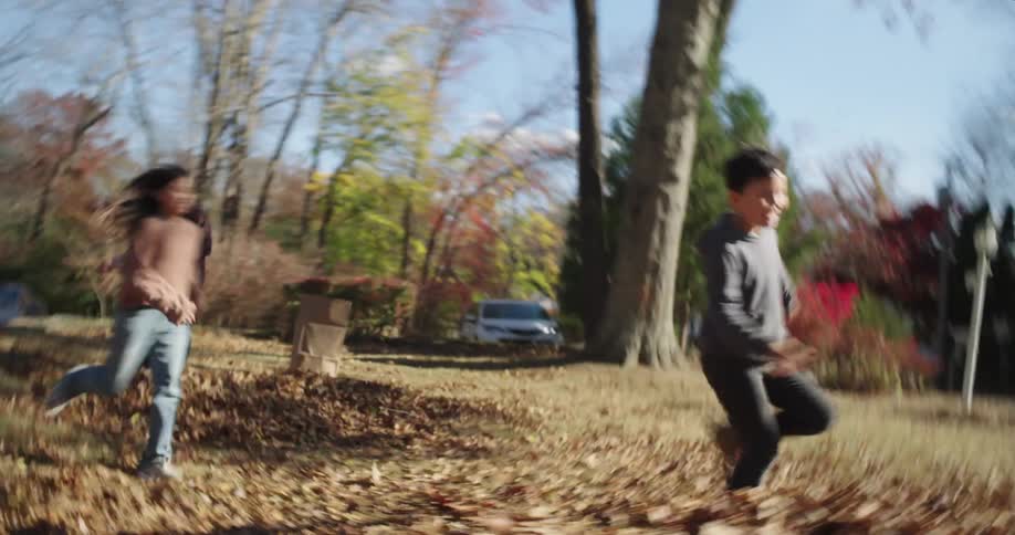 Mother sitting watching the newspaper and two children running out the front door towards the backyard with a dog and playing in the dry leaves fallen on the grass