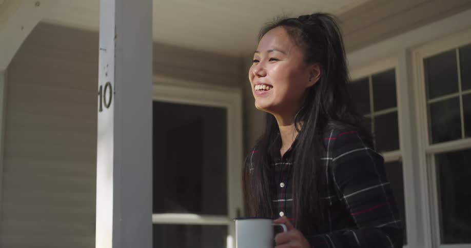 A woman is laughing and talking while holding coffee on her porch