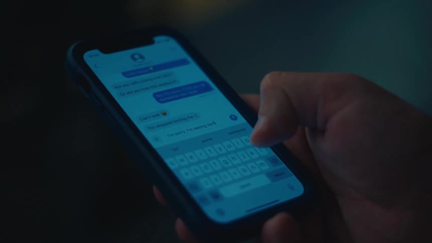 A persons hand is seen typing a message on a smartphone screen illuminated by a blue light