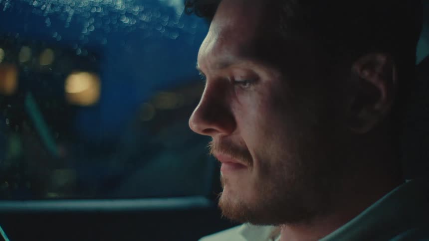 A man sits in a car at night using a smartphone near the rain speckled window illuminated by soft moody lighting