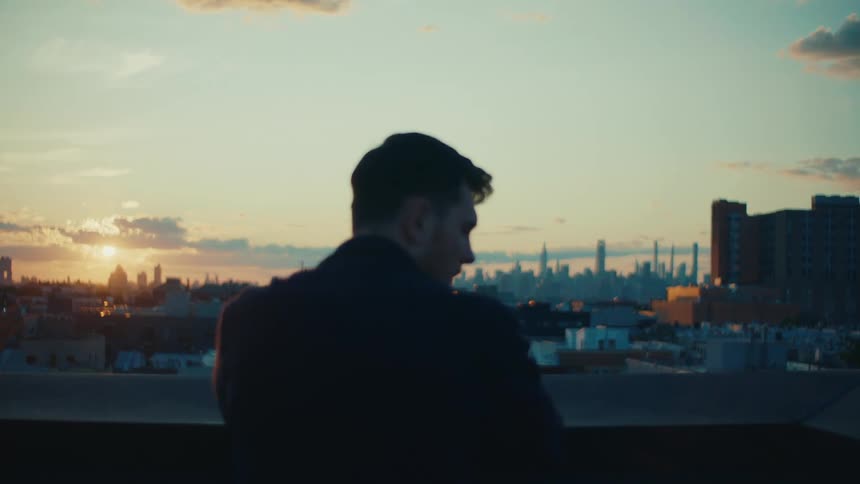 A person is standing on a rooftop at sunset overlooking a city skyline with tall buildings and a colorful sky