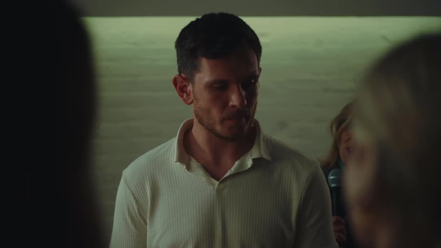 A man with short dark hair wearing a light colored shirt speaks into a microphone in a dimly lit setting