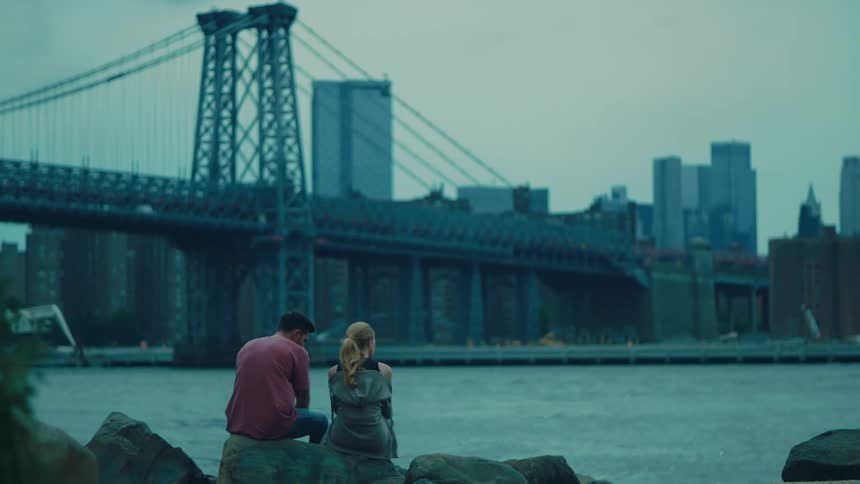 Man and woman sitting together on a rock facing a lake with a view of a metropolis