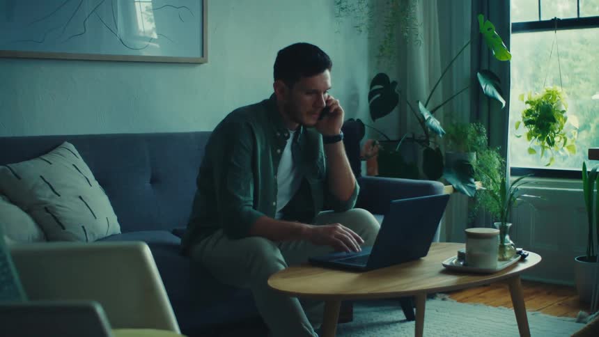 A person is sitting on a couch in a living room talking on the phone while working on a laptop placed on a wooden coffee table