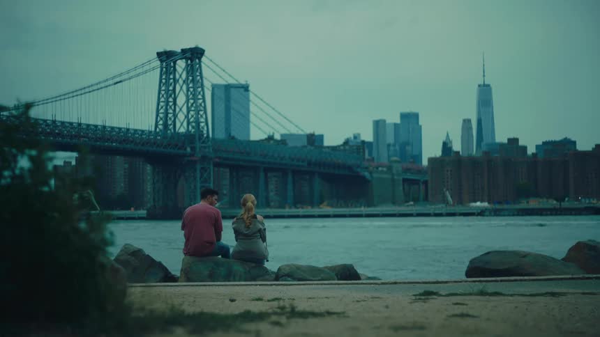View of a man and a woman sitting on a rock by the sea with a bridge and a metropolis in the background