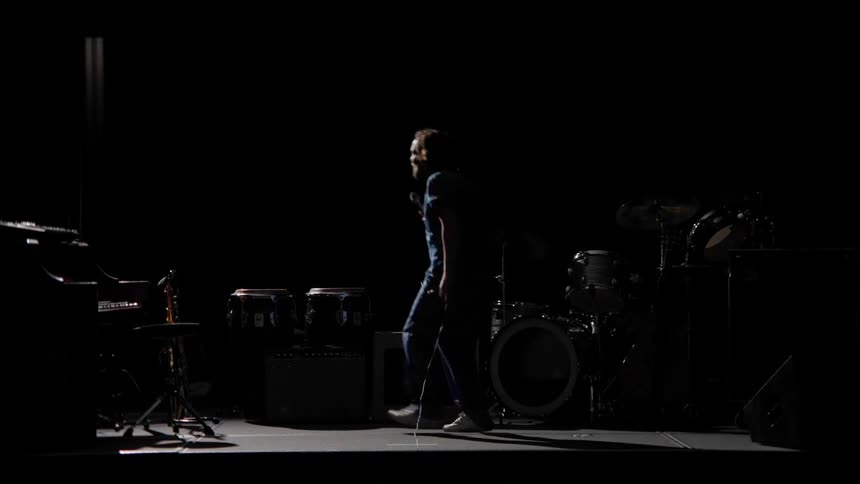 A man holding a microphone hops around a bit on a dark stage while pretending to sing
