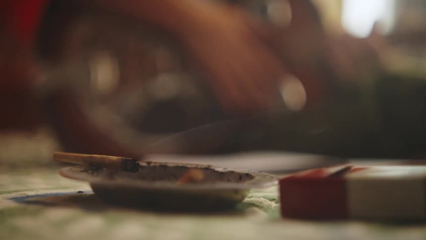 A cigarette that is leaning on top of an ashtray and a little smoke is coming out and behind it there is a person playing the guitar