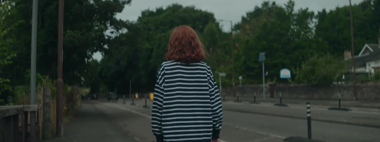 The back of a woman in a striped shirt who is walking on a sidewalk next to an empty asphalt street