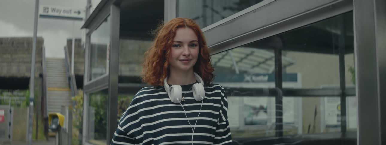 A redhaired woman is wearing a striped shirt and she is standing looking straight ahead with a subtle smile and there is a white headset around her neck