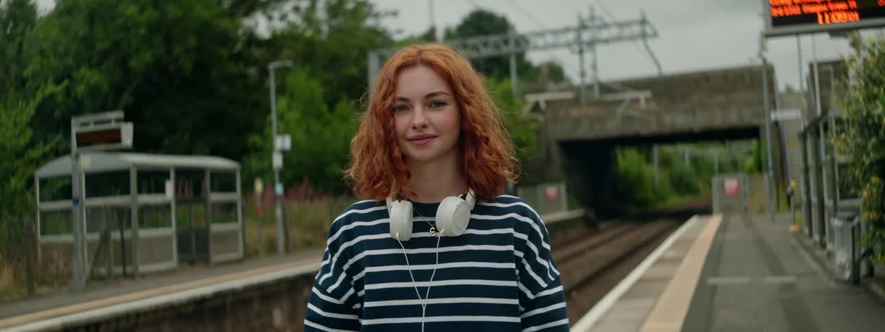 Smiling redheaded woman stands at a train stop near a bridge during the day