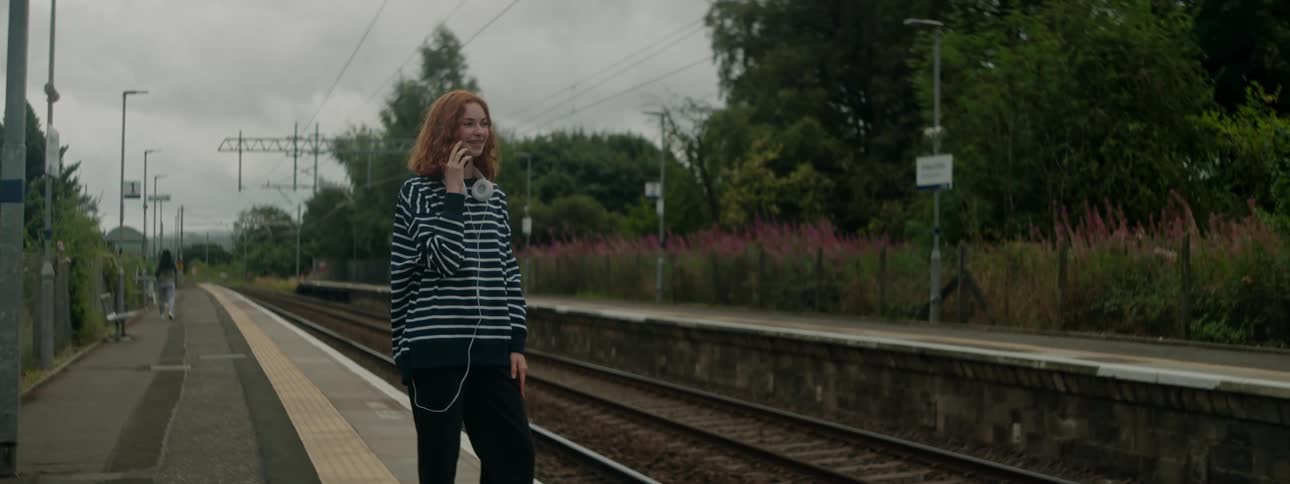 Redheaded woman smiles while talking on a smartphone near the railroad tracks on an overcast day