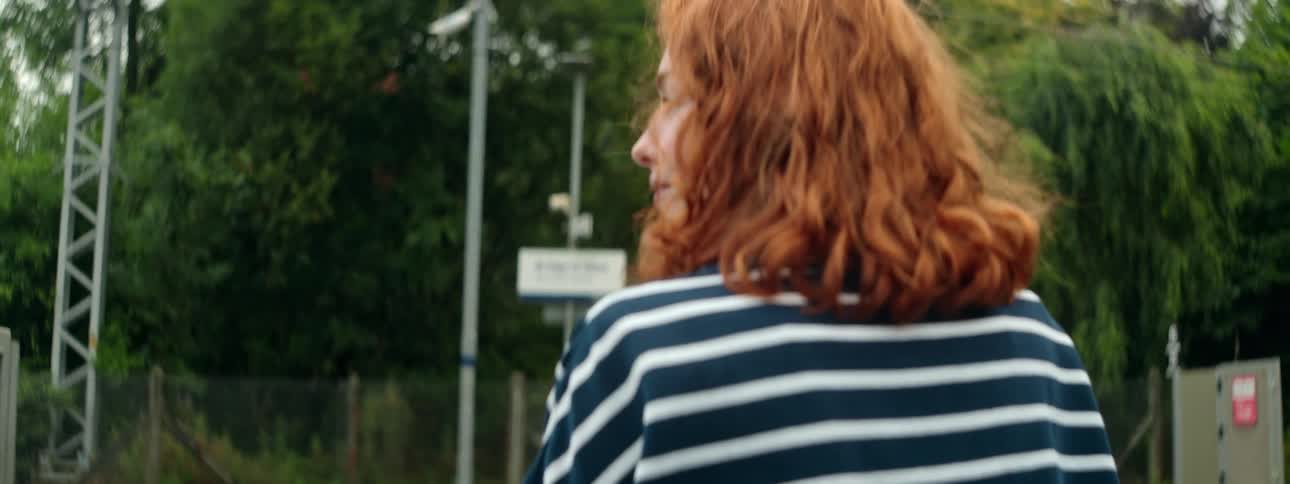 A redhaired woman with a black and white striped shirt and her back is turned looking at a train that is passing by