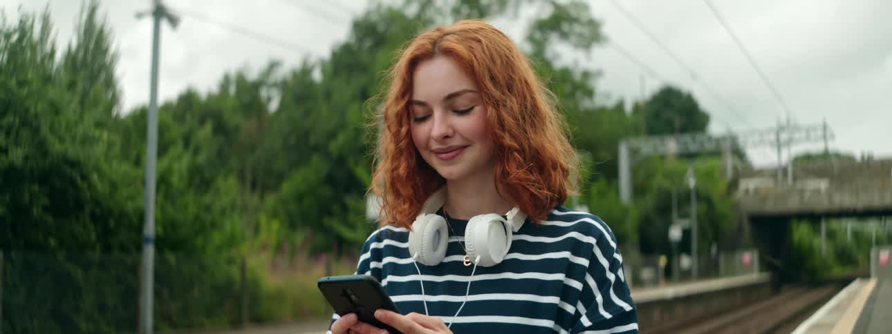 Smiling redhead views a smartphone while walking near the railroad tracks
