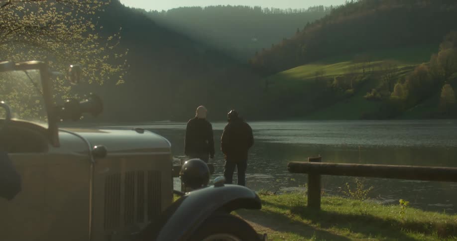 Two men talking by a lake with mountains in the background and a classic car in the foreground