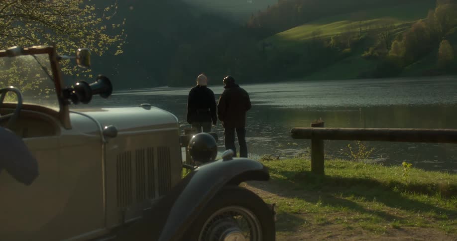 Two people standing on the edge of a large lake are watching it and behind them there is a parked old car