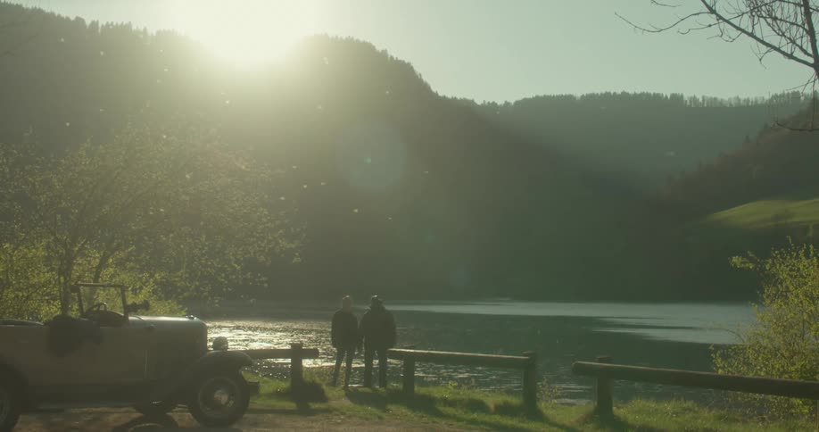 Two figures stand between a lake and an old vintage car silhouetted against the morning sun as it rises over the distant mountains