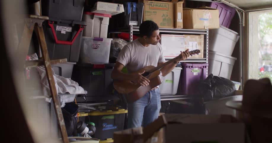 Man picking up a guitar from a garage with boxes and starting to play