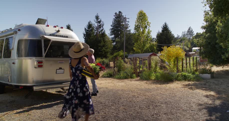 A man cleaning the windows of a trailer while a woman in a dress passes by on her way to a garden