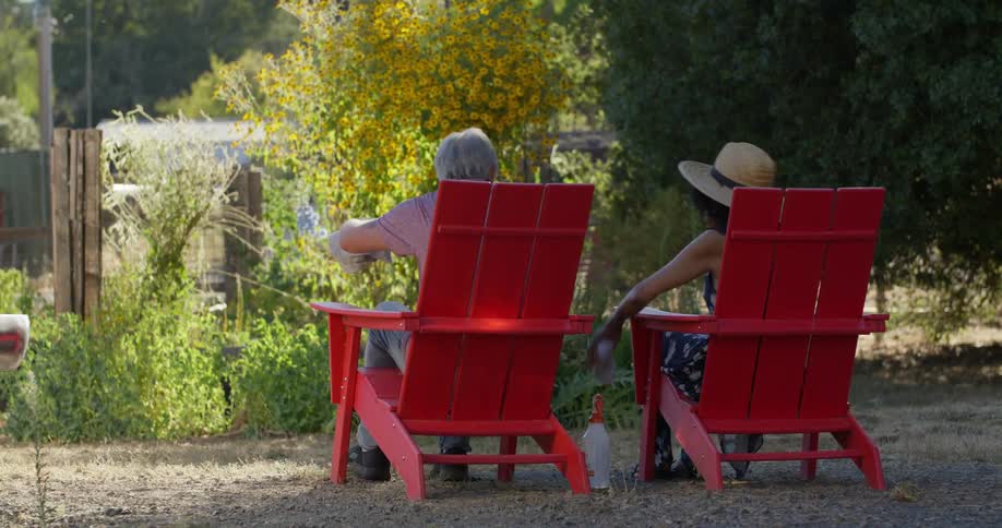 Two people are sitting on bright red adirondack chairs in a garden facing away from the camera