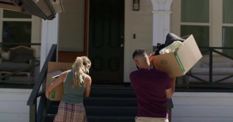 A man and woman unload moving boxes from the back of a vehicle and carry them up the steps to the front door of a house