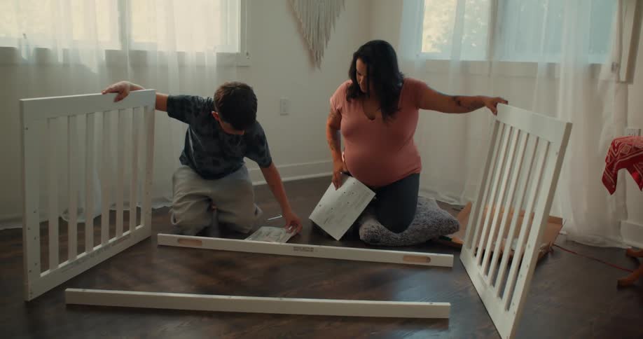 A pregnant woman and her son assemble a crib together in a room