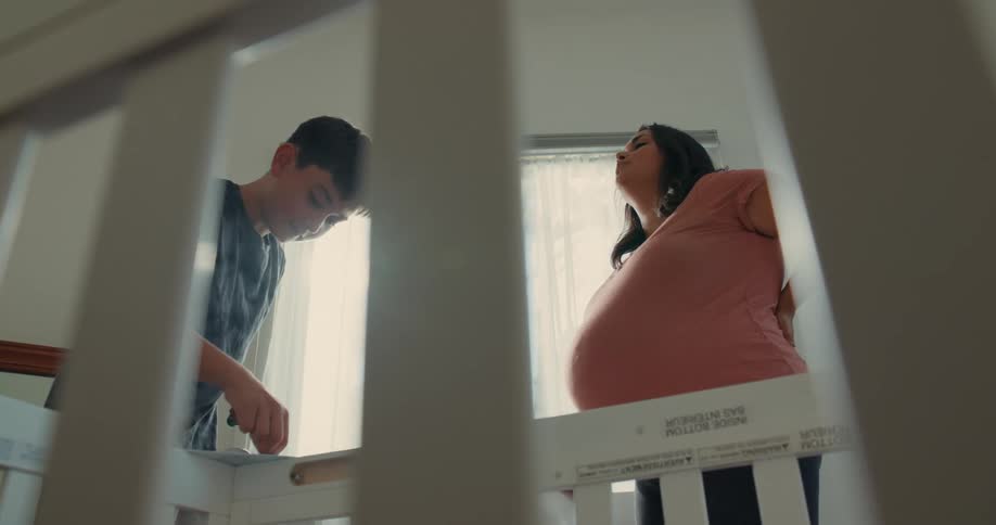 A pregnant mother and her son assembling a white crib in a brightly lit room