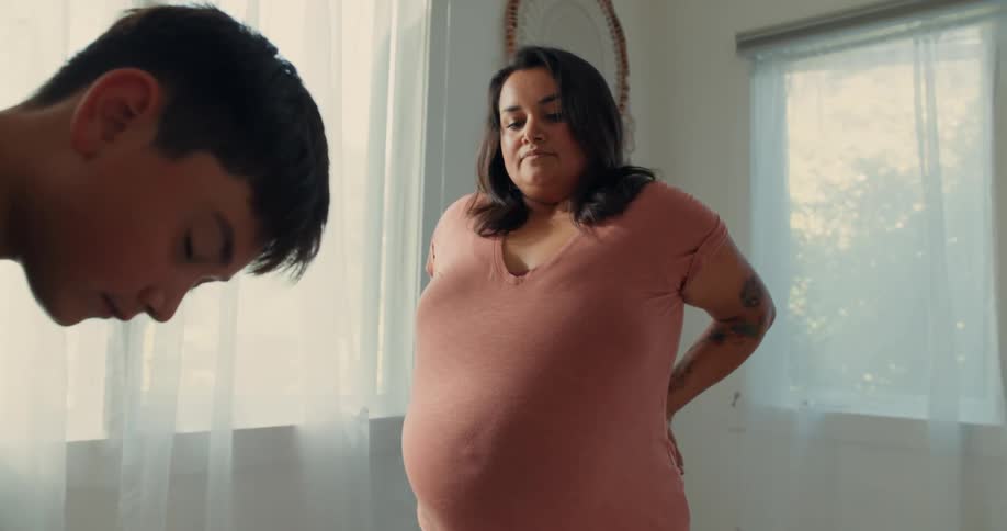 A pregnant mother rubs her belly while her son assembles a crib in a room