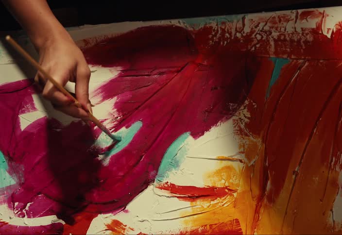 A hand carefully applies a layer of white paint to a vibrant abstract painting with bold strokes of red orange and turquoise