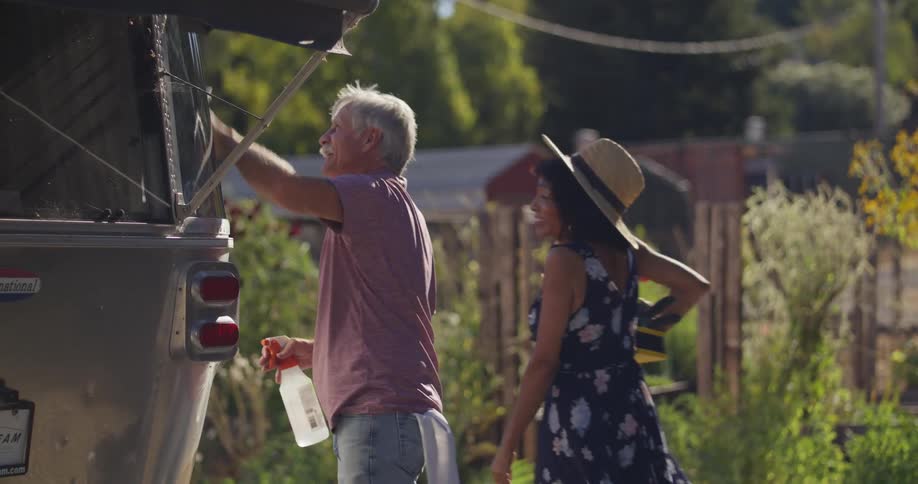An older man with gray hair and a mustache cleans a camper surrounded by a rural setting with lush greenery and rustic buildings in the background while a woman in a blue floral dress carrying a box of flowers walks past