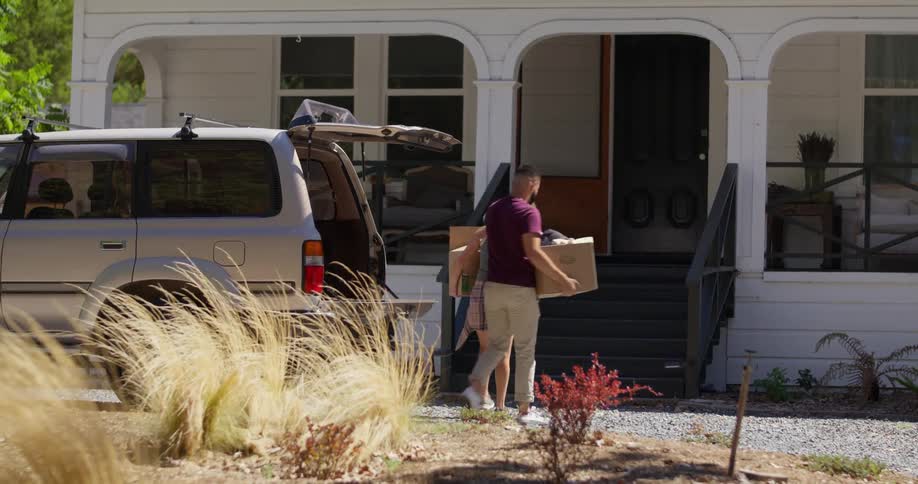 A man and woman carry boxes from the vehicle into the house