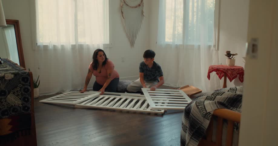 A teenage boy and a woman are sitting on the floor of a room and assembling a white wooden crib