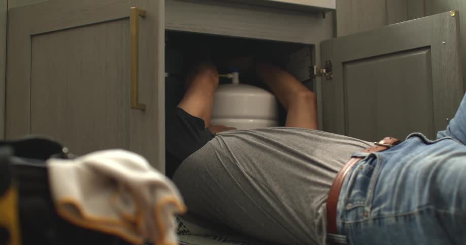 A plumber lying on their back on a rug appearing to hold a wrenchin one hand with tools a bag and a folded cloth nearby suggesting a maintenance or repair scene