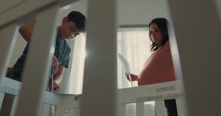 A young boy and a pregnant woman are assembling a crib with the boy focused on tightening something and the woman holding an instruction sheet both framed through the bars of the furniture