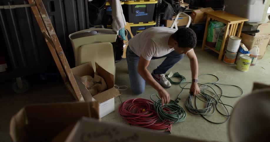 A man in the white shirt and jeans is crouched on the ground and fiddling with a green rope that is coiled
