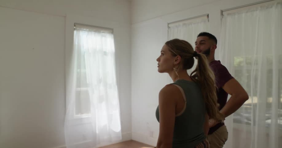 A bearded man in a red shirt and a blonde woman in a green top look around the empty living room of their new house