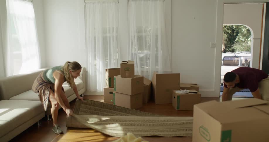 A woman unrolls a rug in a room with moving boxes while a man carries another box inside