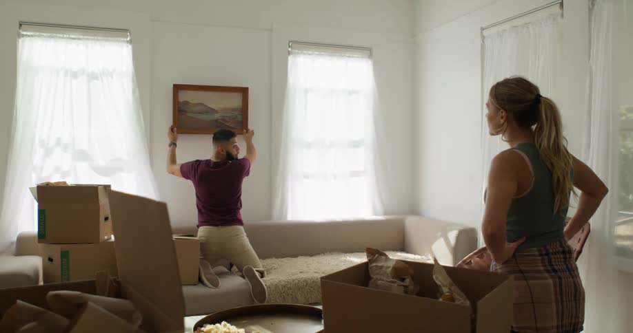 A person is kneeling on a couch hanging a framed picture on the wall while another person stands nearby giving instructions