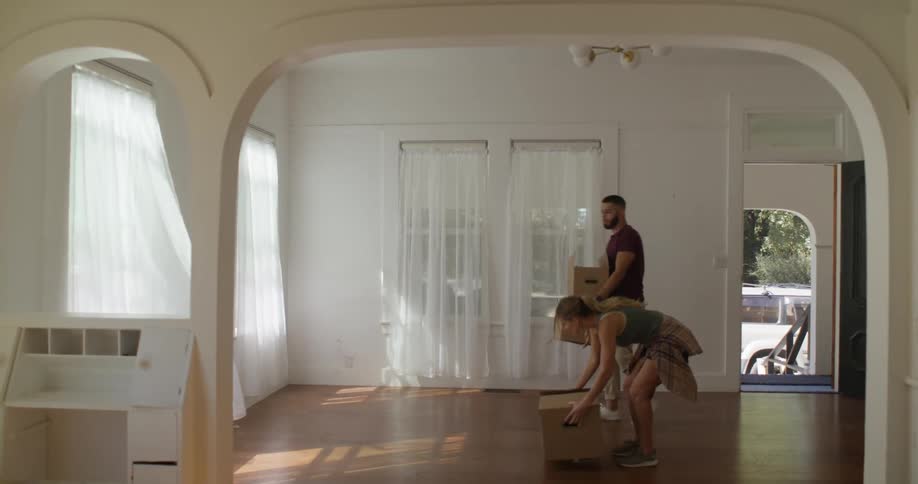 A couple carries boxes into a large empty room with hardwood floors and sheer white curtains