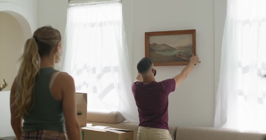 Man adjusting a picture on the wall and a woman behind him adjusting it