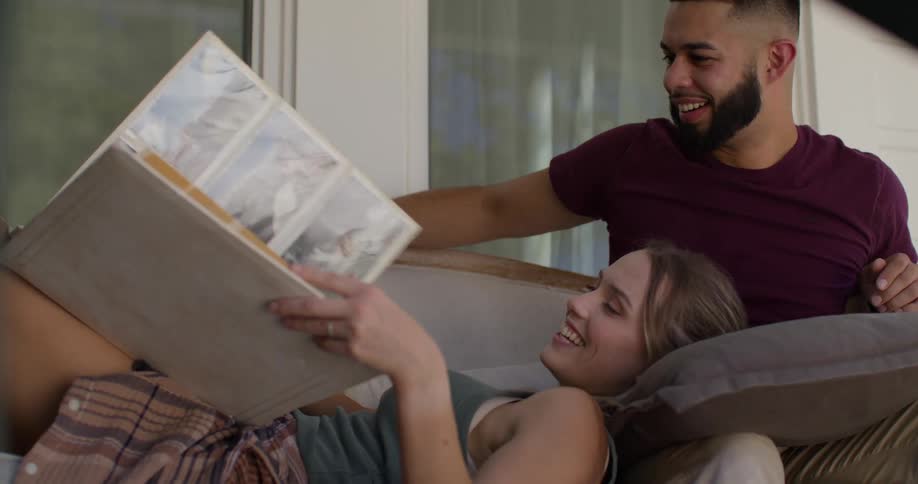 A blonde woman wearing a green top and brown skirt lies on the lap of a bearded man in a red tshirt as she flips through a photo album