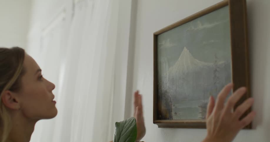 A woman is hanging a painting on the wall of a welllit white room