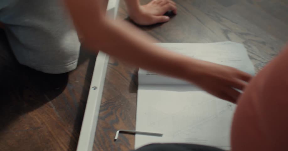 A boy in gray shorts assembles a crib referring to the instruction sheet while a pregnant woman watches nearby