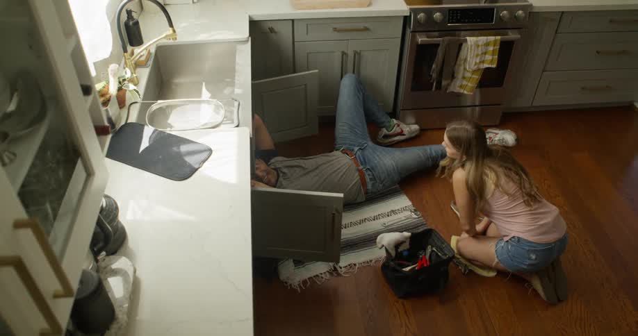 A man lies on the floor working on the plumbing under a kitchen sink while a woman kneels next to him