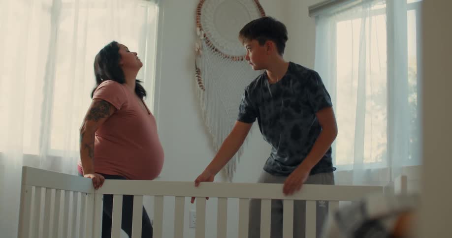 A pregnant woman in a pink tshirt with tattoos on her arm rises from the ground as the boy assembles the crib