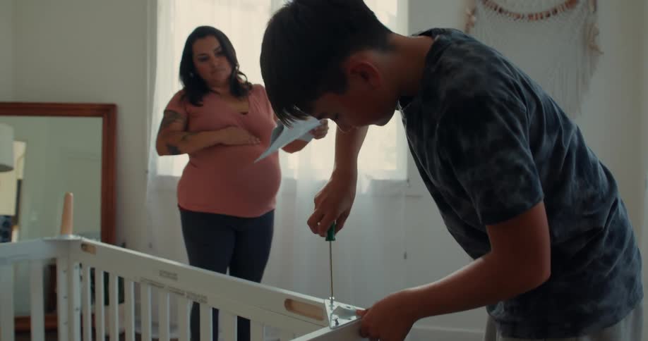 A man is building a crib for a new baby while his pregnant partner watches on holding the instructions