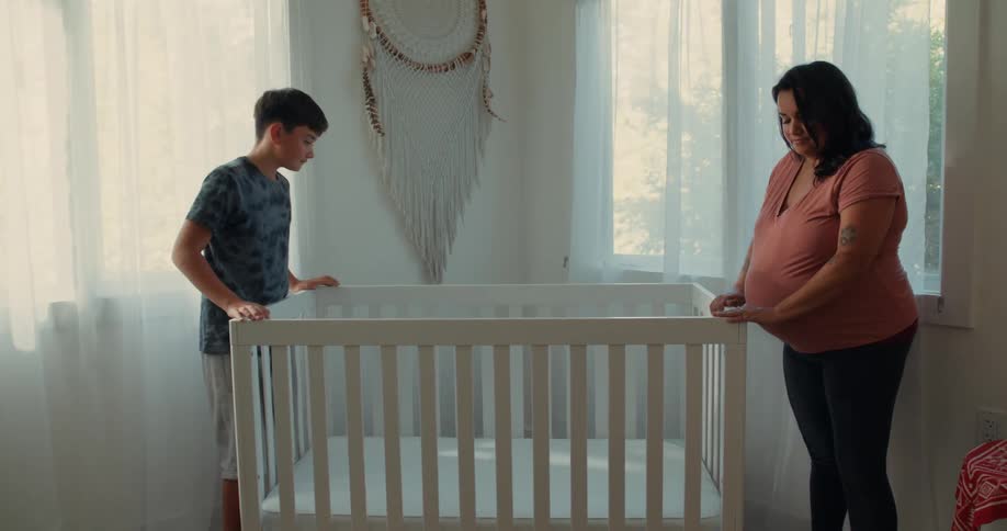 A son and a pregnant mom carrying a crib together in a softly lit room place it down and stand on opposite sides holding each other and gazing down at the crib sharing a heartfelt moment of preparation for their new baby